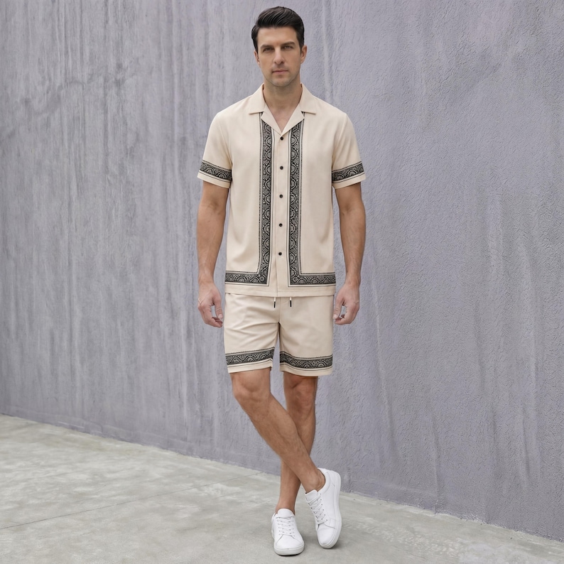 May include: A beige two-piece outfit consisting of a short-sleeved button-up shirt and matching shorts. The shirt and shorts have a black decorative border. The person is wearing white sneakers.