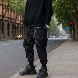 Könnte beinhalten: Schwarze Cargohose mit mehreren Taschen und Riemen, kombiniert mit schwarzen Turnschuhen und einem schwarzen Sweatshirt. Die Hose hat ein modernes, urbanes Design. Die Person steht auf einem Bürgersteig neben einer Straße mit Gebäuden und Bäumen im Hintergrund.