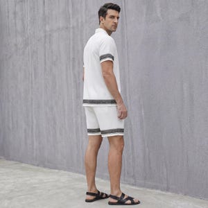 May include: White short-sleeved shirt and matching shorts set with black and gray patterned trim. The shirt has a collared neckline. The shorts have a matching trim at the hem. The model is wearing black sandals.