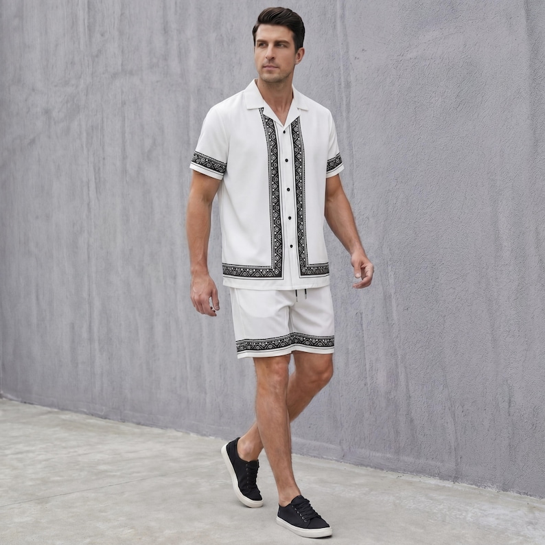May include: A white short-sleeve shirt and matching shorts set. The shirt and shorts have black patterned trim and accents. The shirt has a button-down front. The person is wearing black sneakers.