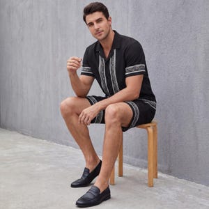 May include: Black short-sleeve shirt and shorts set with white geometric trim. The shirt has a collared neckline and the shorts have an elastic waistband. The model is wearing black leather loafers. The set is made of a lightweight fabric.