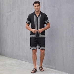 May include: Black two-piece matching set. The short-sleeved shirt and shorts feature white geometric trim. The shirt has a collared neckline and button-up front. The shorts have a matching trim at the hem. The person is wearing brown sandals.
