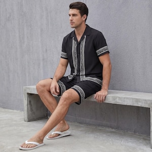 May include: Black short-sleeve shirt and shorts set with white and gray geometric trim. The shirt has a collared neckline and button-down front. The shorts have a matching trim at the hem. White flip-flops complete the outfit.