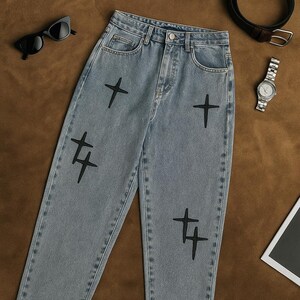 May include: Pale blue denim jeans featuring black cross and stylized plus sign embroidery. The jeans have a classic five-pocket design and a straight leg fit. Accessories include sunglasses, a watch, and a belt.