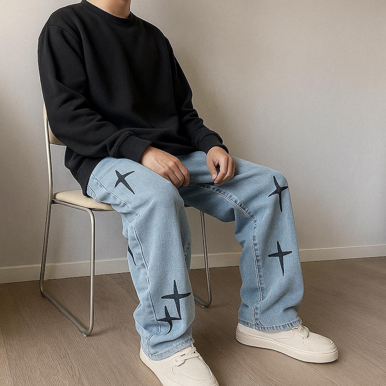 May include: A person wearing a black crew neck sweatshirt and light blue jeans. The jeans feature black star-shaped designs. The person is wearing white trainers and sitting on a beige chair.