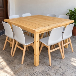 Op de afbeelding: Een grote vierkante eettafel van lichtgekleurd hout met een lattenoppervlak en vier stevige poten, omringd door acht stoelen met witte gevormde zittingen en lichtgekleurde houten poten rondom de tafel geplaatst.