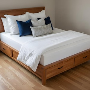 May include: A wooden bed frame with a headboard, two drawers, and a mattress covered with white bedding. The bed is adorned with white and navy blue pillows, and a patterned lumbar pillow. The bed sits on a hardwood floor.