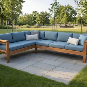 Puede incluir: Un sofá seccional de exterior en forma de L con estructura de madera marrón claro y cojines azules. Varias almohadas decorativas en tonos azules y blancos están dispuestas sobre el sofá. El sofá está colocado en un patio de piedra con un fondo de hierba.