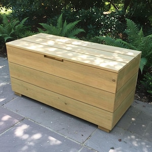 May include: A light-colored wooden storage chest with a rectangular shape. The chest has a slatted top and a recessed handle. It is made of horizontal wooden planks and sits on small feet. The chest is outdoors, surrounded by greenery.