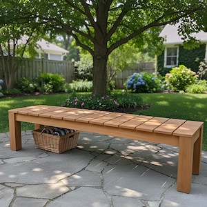 Op de afbeelding: Een houten tuinbank met een lattenbodem en rechthoekige poten. Een rieten mand met blauw-wit gestreepte handdoeken staat onder de bank. De bank staat op een stenen terras.