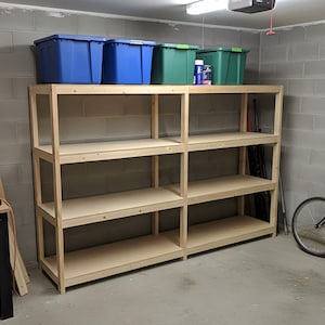 Pode incluir: Uma grande estante de madeira inacabada com quatro prateleiras. Três caixas de armazenamento de plástico azuis e duas verdes estão na prateleira superior. A unidade está em uma garagem com uma bicicleta e ferramentas visíveis.