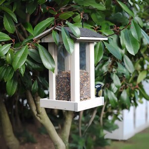 May include: A wooden bird feeder with a black roof and clear panels, filled with birdseed. The feeder is mounted on a tree branch, with a small bird perched on the edge. Green leaves surround the feeder.