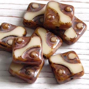 May include: A collection of square-shaped beads featuring a glossy finish with a marbled brown and cream pattern. Each bead displays a distinct curved cream-colored inlay set against a rich brown background with small circular indentations.