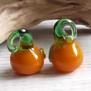 May include: Two small glass pumpkins with orange bodies and green stems. The pumpkins are sitting on a white wooden surface.