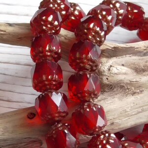 May include: A strand of faceted, deep red beads with a textured, almost floral design. The beads are strung together and rest on a piece of driftwood, creating a contrast of textures and colors. The beads appear to be made of glass or a similar material.