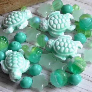 May include: A collection of sea-themed beads in shades of green and blue. The assortment includes turtle-shaped beads, round beads, and star-shaped beads, all with a glossy finish. The beads are arranged on a light-colored wooden surface.