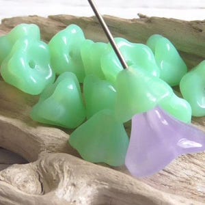 May include: A collection of translucent, light green flower-shaped beads with a single lavender bead. The beads are on a wooden surface, and a metal needle is inserted through one of the beads. These beads are suitable for jewelry making.