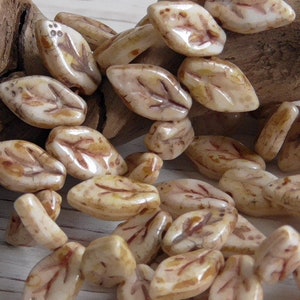 May include: A collection of off-white, leaf-shaped beads with brown vein detailing. The beads have a glossy finish and are scattered, suggesting they are for jewelry making or crafts. The overall color palette is neutral with warm tones.