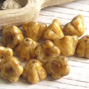 May include: A collection of light brown, flower-shaped beads with a glossy finish. The beads have a detailed, textured surface and a hole for stringing. The background includes a piece of driftwood and a white-painted wooden surface.