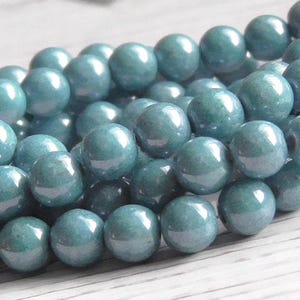 May include: Close-up of a strand of teal-colored, round beads. The beads have a glossy finish and are strung together, creating a textured surface. The beads are approximately 8mm in diameter, suitable for jewelry making.