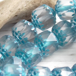 Pode incluir: Um fio de contas de vidro facetadas com um esquema de cores azul claro e turquesa. As contas t&ecirc;m uma superf&iacute;cie texturizada com um padr&atilde;o geom&eacute;trico.