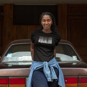 Pode incluir: Uma t-shirt preta de manga curta com a palavra VISUALIZE impressa numa fonte branca repetida, empilhada e desbotada no peito. A peça é usada com calças de ganga azul-escuras e uma camisa de ganga azul-clara atada à cintura.