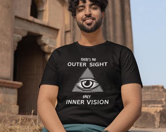 Op de afbeelding: Zwart t-shirt met een witte afbeelding van een oog in een driehoek en de tekst "THERE'S NO OUTER SIGHT ONLY INNER VISION." De persoon draagt een horloge en een teal broek. De achtergrond is een stenen structuur.