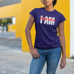 Puede incluir: Una camiseta azul marino con el texto "I AM CERTAIN" en letras blancas con detalles en rojo. La modelo lleva vaqueros azules y gafas. El fondo es una pared amarilla.
