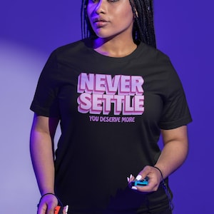 May include: Black t-shirt with the text "NEVER SETTLE YOU DESERVE MORE" in pink and white. The person is holding a red and blue game controller. The background is a gradient of purple and blue.