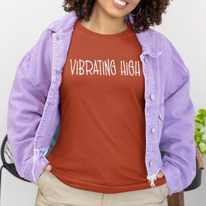 May include: A rust-colored t-shirt with the text "VIBRATING HIGH" in white, worn under a light purple denim jacket. The person is also wearing khaki cargo pants and black sunglasses.