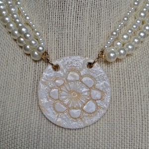 May include: A white ceramic necklace with a round pendant featuring a raised floral design. The pendant is attached to a multi-strand pearl necklace.