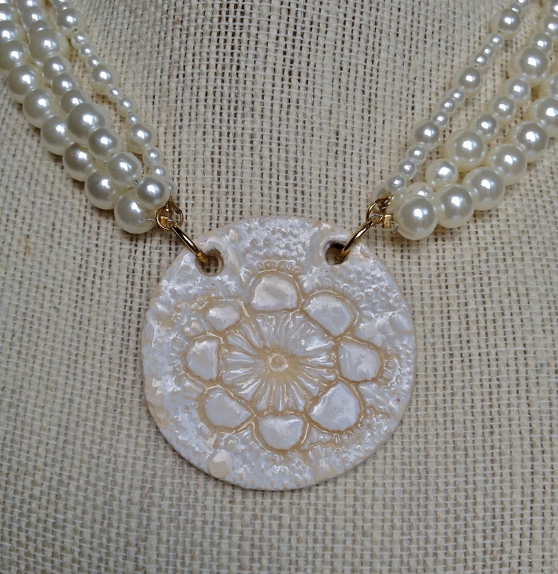 May include: A white ceramic necklace with a round pendant featuring a raised floral design. The pendant is attached to a multi-strand pearl necklace.