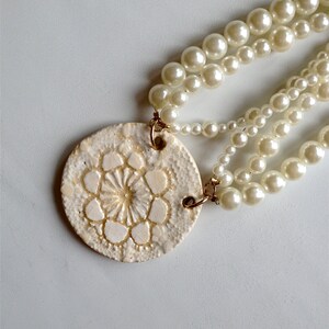 May include: A white ceramic pendant with a floral design hangs from a triple strand of white pearl beads.