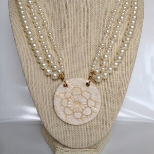May include: A white pearl necklace with a large, white, round pendant featuring a raised, floral design. The pendant is attached to the necklace with gold-colored clasps.