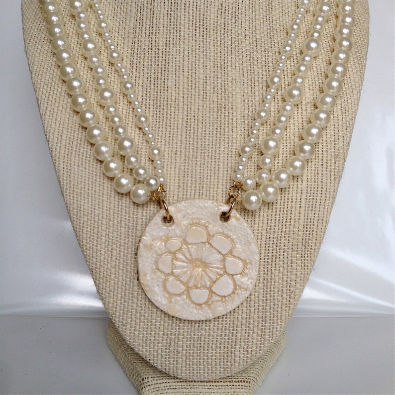 May include: A white pearl necklace with a large, white, round pendant featuring a raised, floral design. The pendant is attached to the necklace with gold-colored clasps.