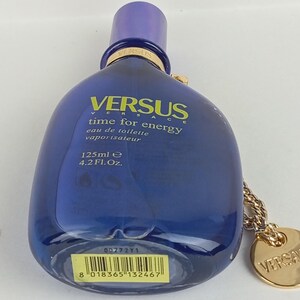 May include: A blue Versus Versace perfume bottle with a purple cap. The bottle has the text "VERSUS time for energy eau de toilette vaporisateur 125ml e 4.2 Fl. Oz." A gold-colored chain and charm are attached.
