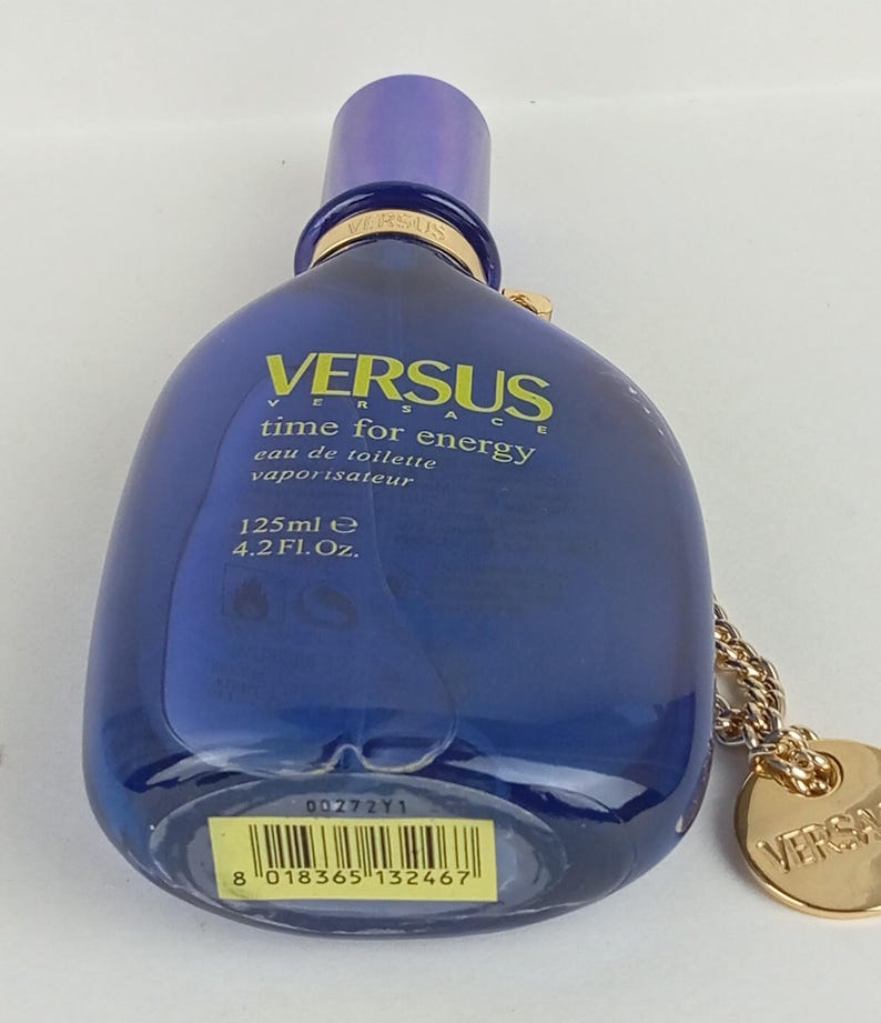 May include: A blue Versus Versace perfume bottle with a purple cap. The bottle has the text "VERSUS time for energy eau de toilette vaporisateur 125ml e 4.2 Fl. Oz." A gold-colored chain and charm are attached.