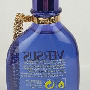 May include: A blue Versus perfume bottle with a gold chain and cap. The bottle has a gold band with the word "VERSUS" and a label with ingredients. The perfume bottle is a stylish fragrance.