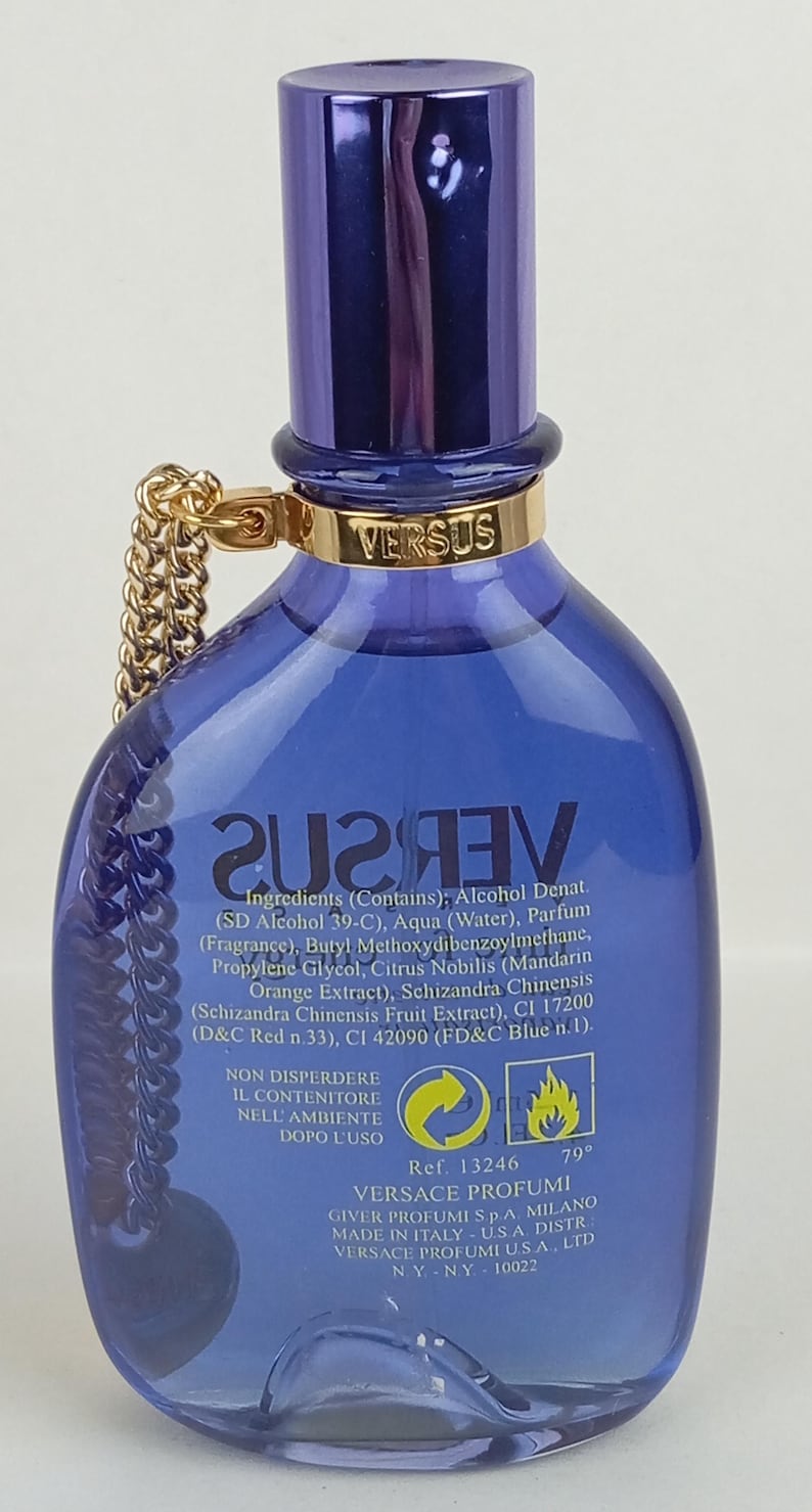 May include: A blue Versus perfume bottle with a gold chain and cap. The bottle has a gold band with the word "VERSUS" and a label with ingredients. The perfume bottle is a stylish fragrance.