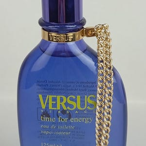 May include: A blue Versus Time for Energy eau de toilette bottle with a gold chain and a gold cap. The bottle has a dark blue cap and a gold band with the word "VERSUS" on it. The bottle contains 125ml or 4.2 Fl. Oz.