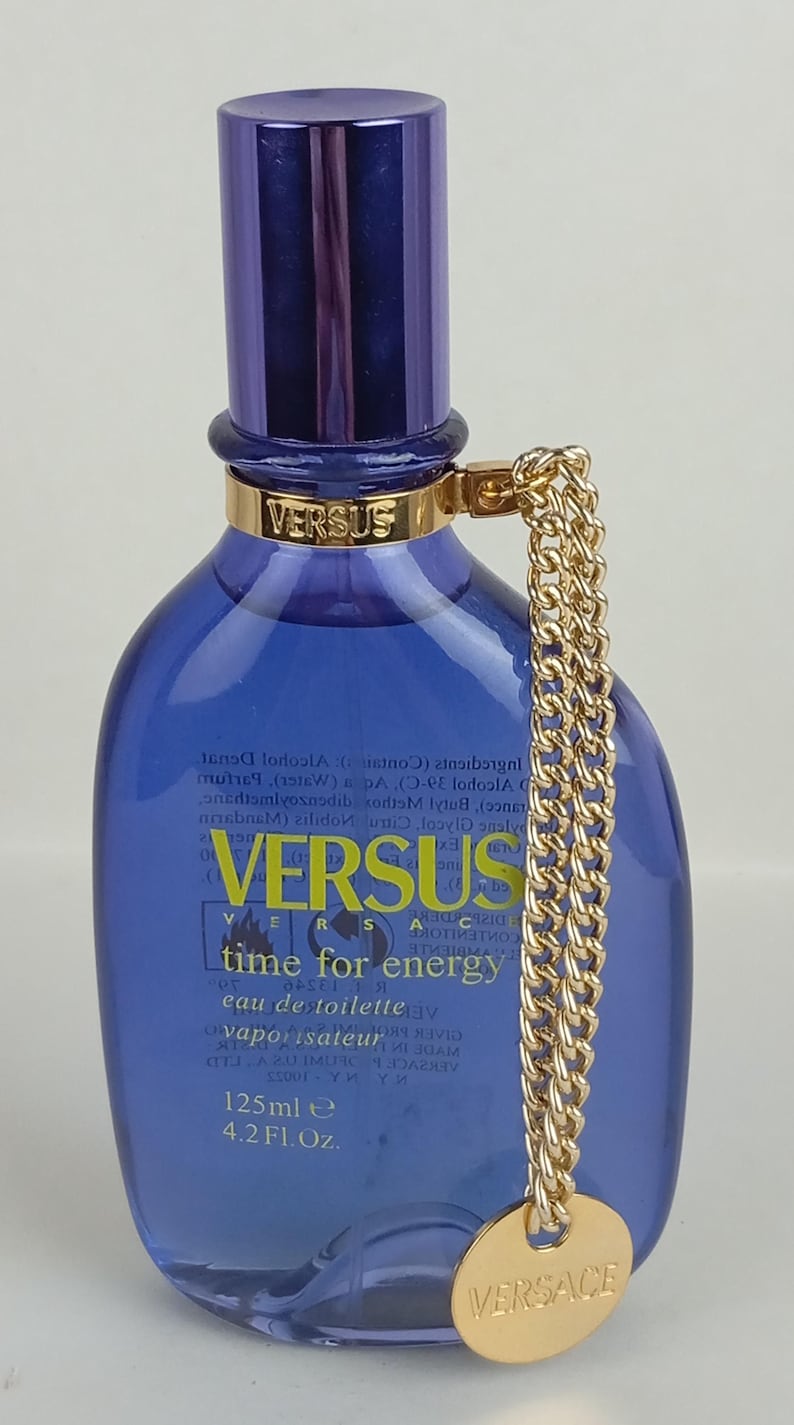May include: A blue Versus Time for Energy eau de toilette bottle with a gold chain and a gold cap. The bottle has a dark blue cap and a gold band with the word "VERSUS" on it. The bottle contains 125ml or 4.2 Fl. Oz.