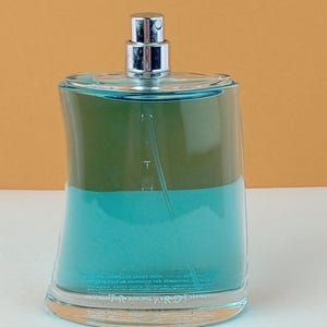 Peut inclure: Un flacon de parfum en verre transparent avec un atomiseur argenté. Le flacon a une forme incurvée unique et est rempli d'un liquide turquoise et vert. Le flacon repose sur une surface blanche sur un fond jaune-orangé.