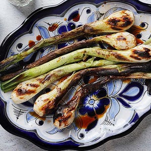 May include: Grilled green onions, charred to perfection, arranged on a decorative, blue and white patterned plate. A halved lemon adds a fresh touch. The onions are a delicious side dish.
