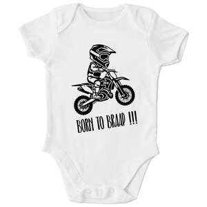 Pode incluir: Um body branco de manga curta com um gráfico preto de uma pessoa usando capacete e pilotando uma moto de motocross. O texto BORN TO BRAAP !!! está impresso em preto abaixo do gráfico, que também inclui as letras MX no quadro da moto.