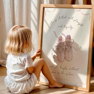 Puede incluir: Impresión enmarcada con un par de zapatillas de ballet rosas y el texto «Ella está vestida de fuerza y dignidad, y se ríe sin temor al futuro. Proverbios 31:25». El marco es de madera clara.