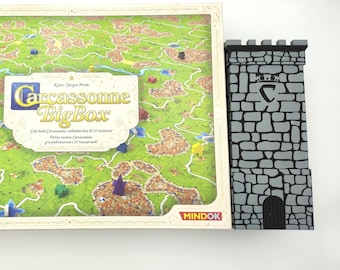 May include: A Carcassonne BigBox board game box featuring a green landscape illustration with small colored wooden figures and a separate grey stone-textured tower accessory with a black door and a crown emblem printed on the front surface.