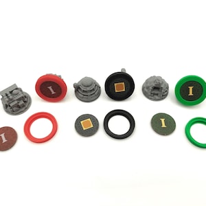 May include: A collection of game pieces, including gray miniature structures, red and green rings, and circular tokens with the letter "I". The pieces are in various colors, including red, green, black, and brown, and are arranged on a white surface.