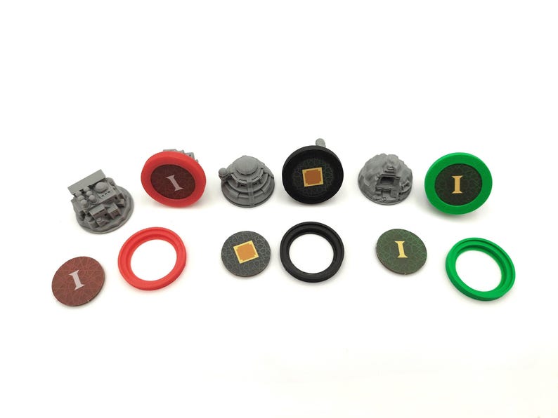May include: A collection of game pieces, including gray miniature structures, red and green rings, and circular tokens with the letter "I". The pieces are in various colors, including red, green, black, and brown, and are arranged on a white surface.
