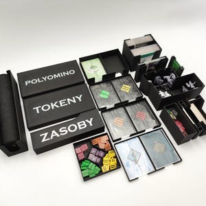 May include: A collection of black game component boxes and trays. The boxes are labeled "POLYMINO", "TOKENY", and "ZASOBY" in white text. Various game pieces, cards, and miniatures are visible within the containers, suggesting a board game organization system.