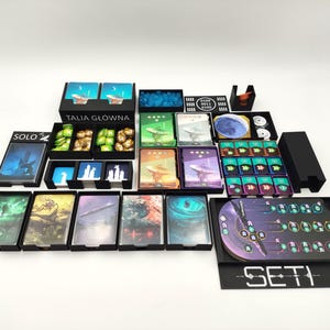 May include: A collection of board game components, including cards, tokens, and storage boxes. The set features various colors and designs, with text that reads "TALIA GŁÓWNA", "SOLO", and "SETI".